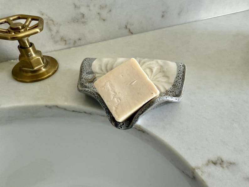 May include: A square bar of light-colored soap rests in a decorative soap dish. The dish has a gray and white floral design and a textured rim. A brass faucet is visible in the background, suggesting a bathroom setting.