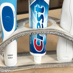 May include: A white ceramic toothbrush holder with three slots. The holder has a speckled glaze and a gray rim. There are two toothbrushes and a tube of Crest toothpaste in the holder.