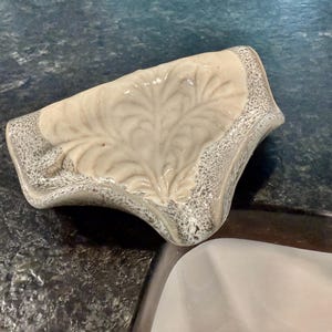 May include: A ceramic soap dish with a leaf design. The dish is off-white with a gray speckled border. The leaf pattern is raised and detailed. The dish is curved and sits on a dark countertop.