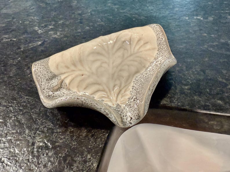May include: A ceramic soap dish with a leaf design. The dish is off-white with a gray speckled border. The leaf pattern is raised and detailed. The dish is curved and sits on a dark countertop.