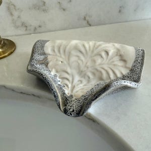 May include: A ceramic soap dish with a white floral design and a speckled gray border. The dish is placed on a white marble countertop near a brass faucet, showcasing a blend of functionality and decorative appeal.