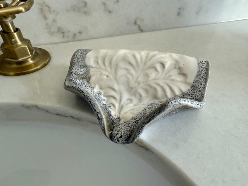 May include: A ceramic soap dish with a white floral design and a speckled gray border. The dish is placed on a white marble countertop near a brass faucet, showcasing a blend of functionality and decorative appeal.