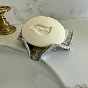 May include: A round, off-white bar of soap with the word "source" and a leaf design is in a gray, textured soap dish. A brass faucet is in the background. The soap dish has a wavy, organic shape.