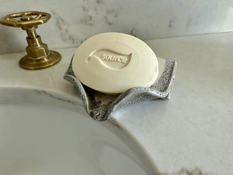 May include: A round, off-white bar of soap with the word "source" and a leaf design is in a gray, textured soap dish. A brass faucet is in the background. The soap dish has a wavy, organic shape.
