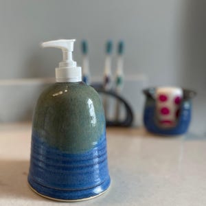 May include: A ceramic soap dispenser with a white pump top. The dispenser is a mix of blue and green hues, with a ribbed design at the base. A matching blue ceramic holder with a pink and white item is in the background.
