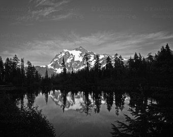 May include: Black and white landscape photograph of a mountain range with snow-capped peaks reflected in a calm lake. Dark silhouettes of evergreen trees line the water's edge, with a cloudy sky overhead.