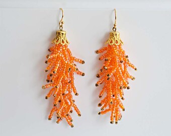 May include: Orange beaded tassel earrings with gold metal tops. The earrings are approximately 7.5 cm long.