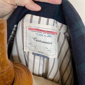 May include: Close-up of a dark blue and green plaid jacket interior. The jacket lining is striped with tan and white. A label reads "Cartonnier" with text above it that says "Conservation and Restoration".