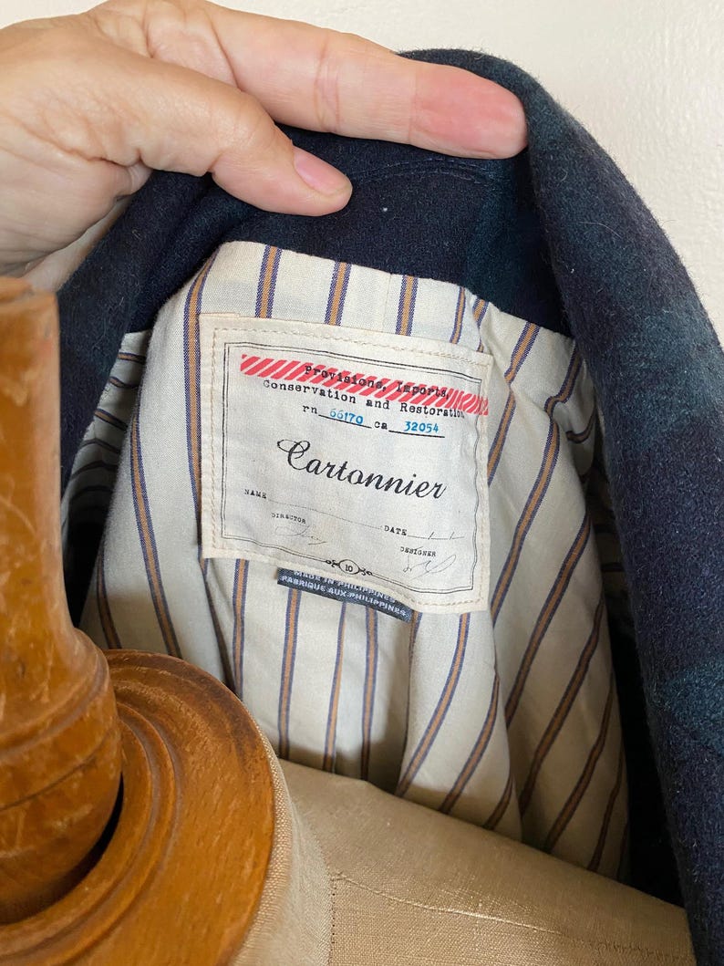 May include: Close-up of a dark blue and green plaid jacket interior. The jacket lining is striped with tan and white. A label reads "Cartonnier" with text above it that says "Conservation and Restoration".