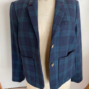 May include: A navy blue and teal plaid blazer with two front pockets and two buttons. The blazer is displayed on a mannequin. The blazer has a classic design.