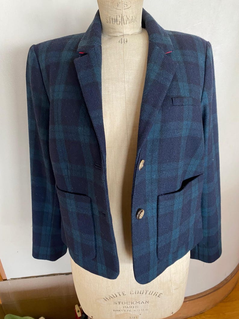 May include: A navy blue and teal plaid blazer with two front pockets and two buttons. The blazer is displayed on a mannequin. The blazer has a classic design.