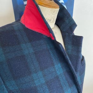 May include: A close-up of a plaid wool coat with a red collar. The coat features a navy blue and teal plaid pattern. A gold button is visible on the front. The coat is displayed on a mannequin.