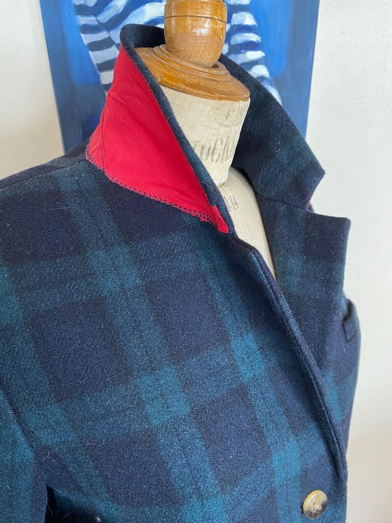 May include: A close-up of a plaid wool coat with a red collar. The coat features a navy blue and teal plaid pattern. A gold button is visible on the front. The coat is displayed on a mannequin.