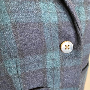 May include: Close-up of a navy and teal plaid blazer. The blazer features two gold-colored buttons and a small pocket. The fabric has a textured appearance.