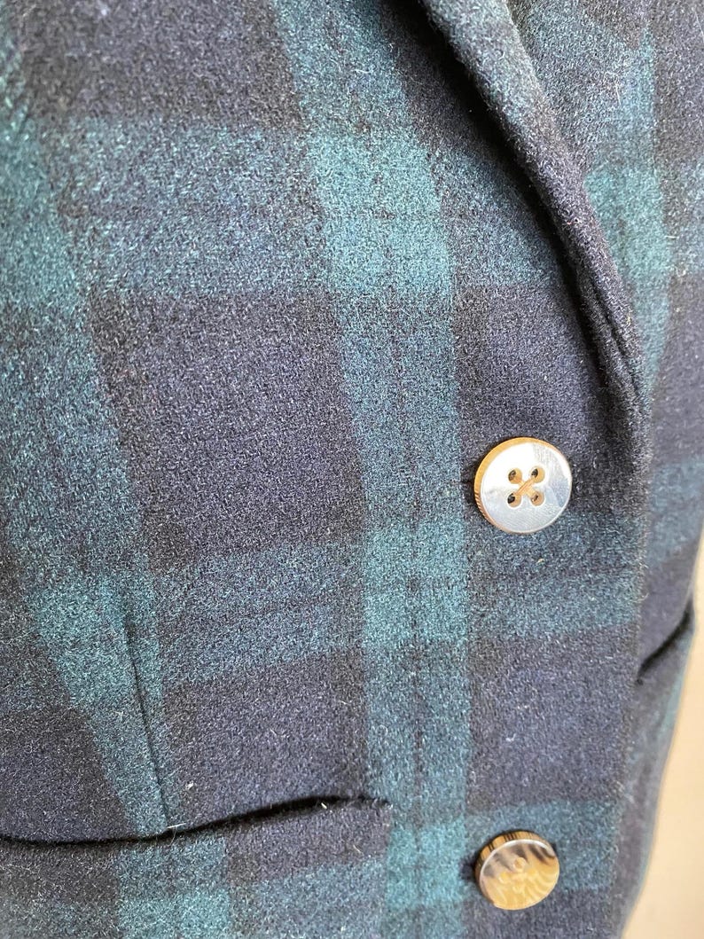 May include: Close-up of a navy and teal plaid blazer. The blazer features two gold-colored buttons and a small pocket. The fabric has a textured appearance.