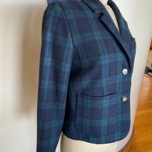 May include: A navy blue and teal plaid blazer with two decorative buttons and a single pocket. The blazer is displayed on a mannequin. The blazer has a classic, tailored design.
