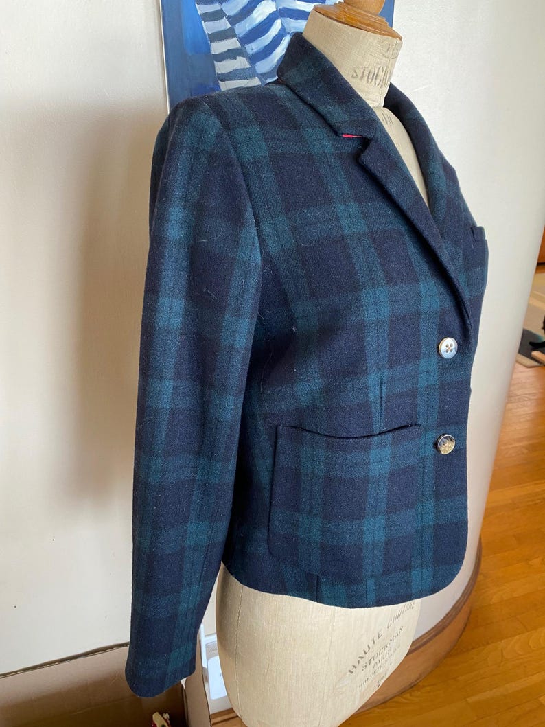 May include: A navy blue and teal plaid blazer with two decorative buttons and a single pocket. The blazer is displayed on a mannequin. The blazer has a classic, tailored design.