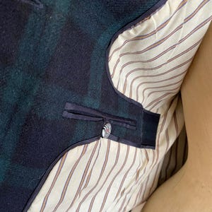 May include: Close-up of a dark green and navy blue plaid blazer. The interior lining is cream-colored with thin brown and orange stripes. A single button with a black, white, and red design is visible.