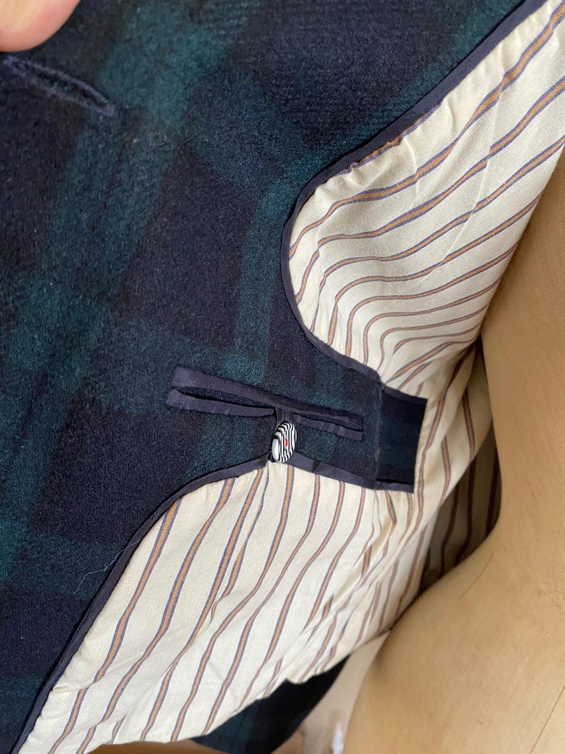 May include: Close-up of a dark green and navy blue plaid blazer. The interior lining is cream-colored with thin brown and orange stripes. A single button with a black, white, and red design is visible.