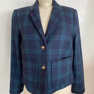 May include: A navy blue and teal plaid blazer with two front pockets and two gold-colored buttons. The blazer is displayed on a mannequin. The blazer has a notched lapel and a red stitch detail on the collar.