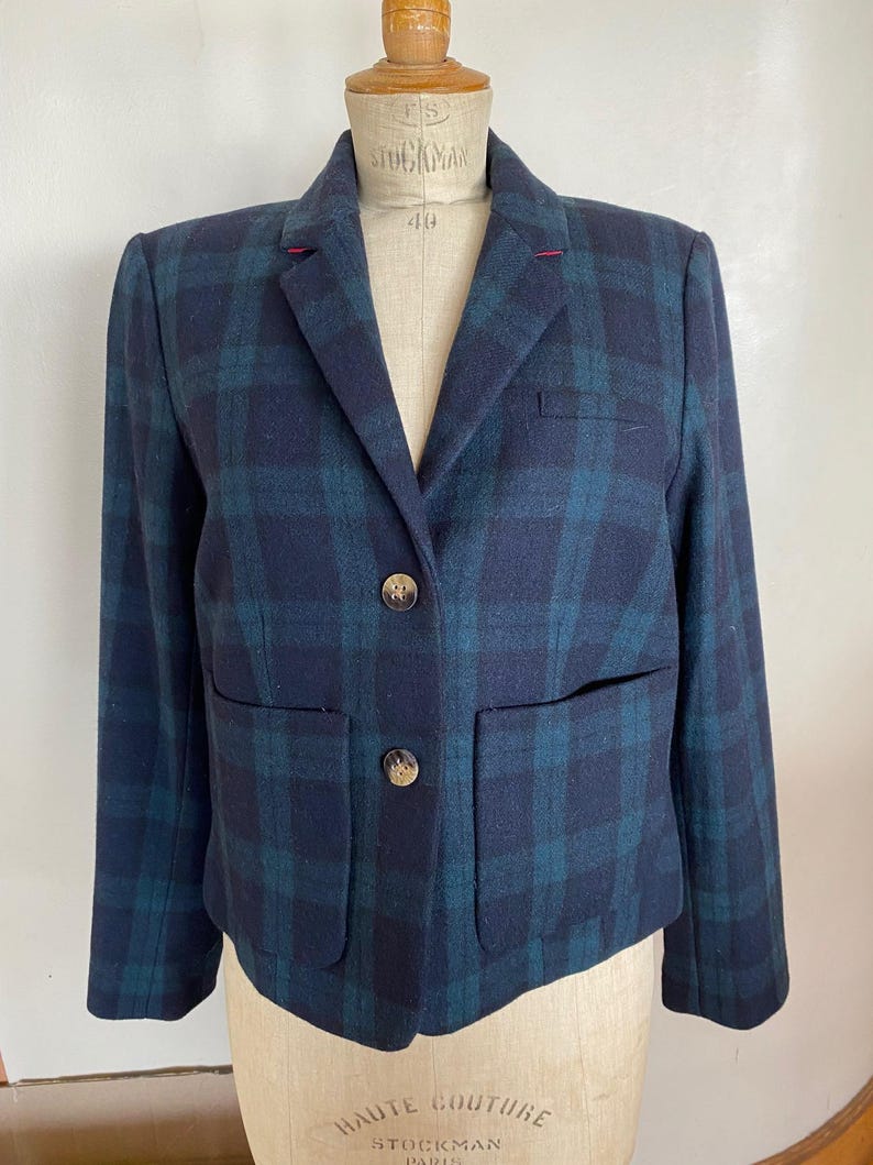 May include: A navy blue and teal plaid blazer with two front pockets and two gold-colored buttons. The blazer is displayed on a mannequin. The blazer has a notched lapel and a red stitch detail on the collar.