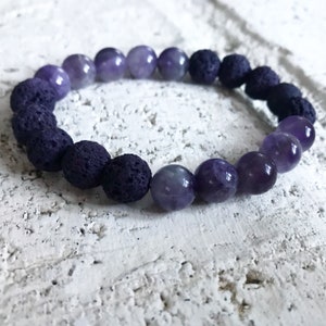 May include: A purple amethyst and black lava rock beaded bracelet. The bracelet is made with round beads and has a gradient of purple amethyst beads.