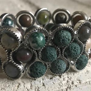 Op de afbeelding: Een zilveren armband met een elastische band heeft een verscheidenheid aan groene, bruine en grijze stenen. De stenen zijn gezet in zilveren settings en zijn gerangschikt in een cirkelvormig patroon.