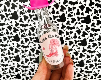 May include: A small glass bottle with a pink cowboy hat on top. The bottle has a pink and white label with the text "Let's Go Girls" and "Ella's Last Rodeo 2023".