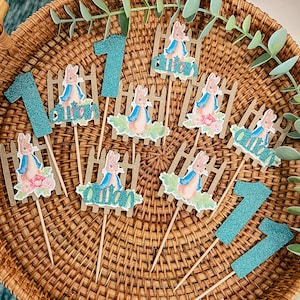 May include: A collection of Peter Rabbit themed cupcake toppers and teal glitter number one toppers arranged on a woven basket. The toppers feature Peter Rabbit illustrations, picket fences, and the name "auton".