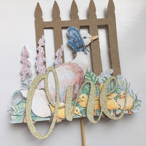 May include: A gold glitter cake topper featuring a duck with a blue bonnet and her ducklings. The duck is standing in front of a brown picket fence with pink and blue flowers. The word "one" is written in gold glitter.