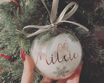 May include: A clear glass Christmas bauble with a white feather inside and the name "Millie" written in rose gold lettering. The bauble is hanging from a grey ribbon.