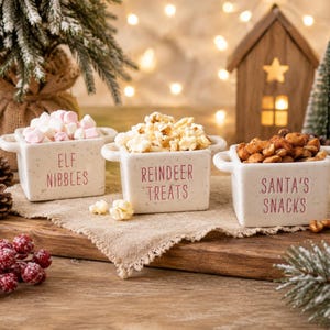 May include: Three square white ceramic bowls with side handles and speckled texture. Each bowl features red text: one reads ELF NIBBLES filled with marshmallows, one reads REINDEER TREATS filled with popcorn, and one reads SANTA'S SNACKS filled with nuts.