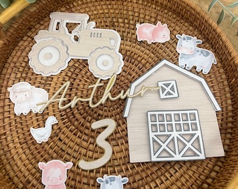 May include: A selection of farm animal cake toppers, featuring a tractor, barn, sheep, goose, pigs, and cows. The name "Arthur" is written in gold script, and the number "3" is included. The toppers are displayed on a woven wicker tray.