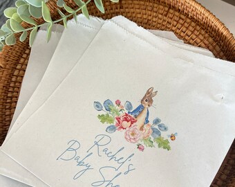 May include: White paper gift bags with a Peter Rabbit illustration and the text "Rachel's Baby Shower" printed in blue.