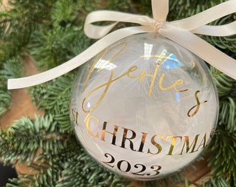 May include: A clear glass Christmas bauble with a white ribbon and gold lettering. The bauble says "Ellie's First Christmas 2023" and has white feathers inside.