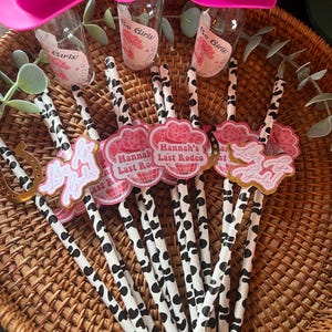May include: A set of pink cowboy hats with a cow print straw and a pink and gold party favor with the text "Hannah's Last Rodeo" on it.