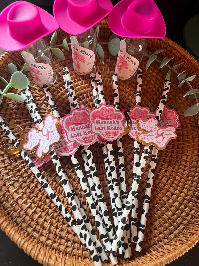 May include: A set of pink cowboy hats with a cow print straw and a pink and gold party favor with the text "Hannah's Last Rodeo" on it.