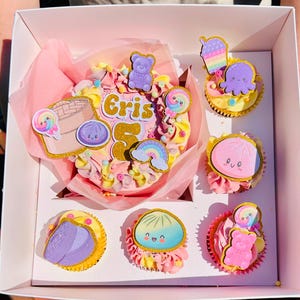 May include: A round cake with pink and yellow frosting, topped with gold glitter cutouts of a bear, a rainbow, a lollipop, a dumpling, and the text Eris 5, accompanied by five cupcakes featuring similar colorful character and food toppers.