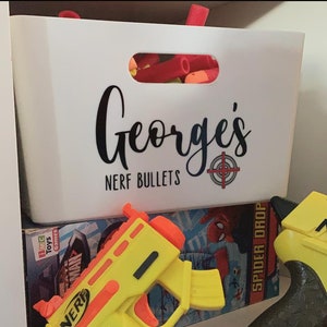 Puede incluir: Un contenedor de almacenamiento de plástico blanco con el nombre "George's" y las palabras "NERF BULLETS" impresas en la parte delantera. El contenedor está lleno de dardos NERF rojos y naranjas. Una pistola NERF amarilla está en primer plano.
