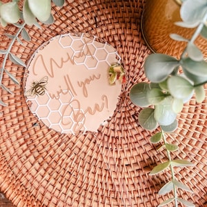 May include: A round, clear acrylic sign with a beige background and white honeycomb pattern. The text "What will you Bee?" is written in gold script. A small gold bee is on the left side of the sign and a gold honeycomb is on the right side.