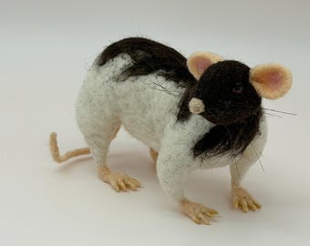 May include: A needle-felted rat figurine featuring a black head and a white body with a black patch along the back. The rat has large pink-lined ears, a small cream-colored nose, dark eyes, thin whiskers, and light-colored paws and tail.