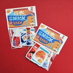 Op de afbeelding: Drie pakjes Japanse snackstickers op een rode achtergrond. Elk pakje bevat een kaart met de woorden "SNACK PACK" en Japanse karakters. De stickers tonen kleurrijke illustraties van Japanse snacks en drankjes, waaronder een melkpak.