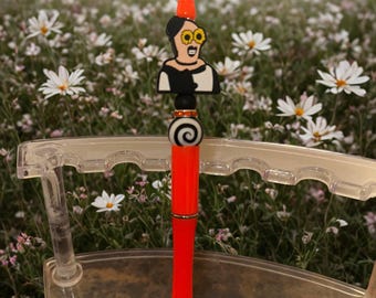 May include: A bright red pen with a decorative topper featuring a cartoon woman with yellow glasses and a black and white outfit. The pen is displayed in a clear plastic pen holder, set against a backdrop of white flowers.