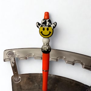 May include: A novelty pen with a bright orange barrel and a smiley face charm wearing a cowboy hat. The pen has a silver tip and a decorative bead section with a black and silver glitter ball.