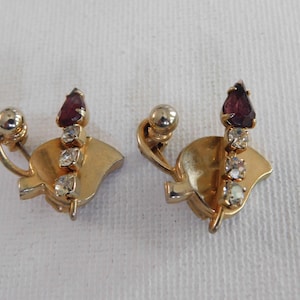 May include: A pair of gold tone clip-on earrings with a leaf design. Each earring features a single purple rhinestone and four clear rhinestones.