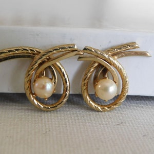 May include: A pair of gold-toned clip-on earrings. Each earring features a faux pearl suspended within a swirling, textured gold design. The earrings have a bow-like shape with a textured, rope-like pattern on the gold metal.