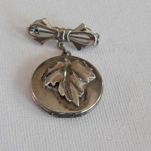 May include: A silver locket with a detailed leaf design on the front. The locket is attached to a silver bow pin.