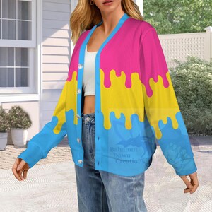 Op de afbeelding: Een vest met lange mouwen en knopen met een levendig driekleurig ontwerp met horizontale banden van roze, geel en blauw. De kleuren zijn gerangschikt in een druipend verfpatroon. Het vest heeft een lichtblauwe rand langs de opening.