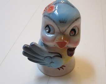 May include: A blue ceramic bird figurine with a yellow beak, pink flowers, and sparkling rhinestone eyes. The bird has a blue and gray wing and is standing on a white base.