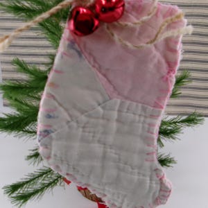 May include: A pink and white patchwork fabric stocking ornament with red jingle bells attached to the top.